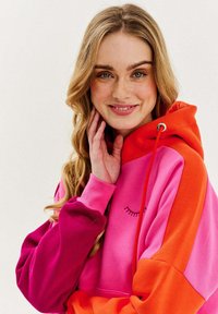 Barevná mikina s velkými panely v růžové, fuchsiové a oranžové barvě. Má kapuci se stahovací šňůrkou a malou grafiku oka na hrudi. Měkký materiál.