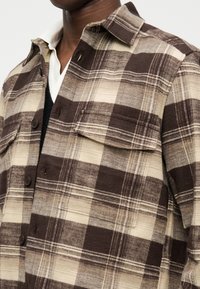 Nahaufnahme einer Person, die ein braun-beiges kariertes Hemd über einem weißen Hemd mit Kragen und einem dunklen Pullover trägt.