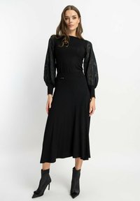 Czarna sukienka z długimi, bufkowymi koronkowymi rękawami i dopasowanym gorsetem, charakteryzująca się gładką fakturą i kokardą w talii. Stylizowana z czarnymi botkami za kostkę.