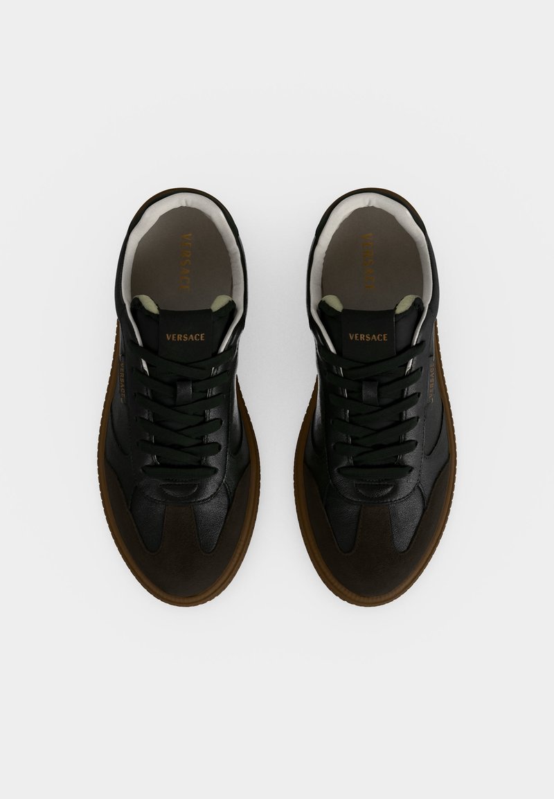 Coppia di sneaker Versace in pelle nera con suole marroni e lacci neri, vista dall'alto su sfondo bianco.