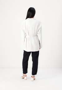 Giacca oversized bianca texturizzata con cintura, abbinata a pantaloni neri larghi e sandali bianchi con tacco a blocco.