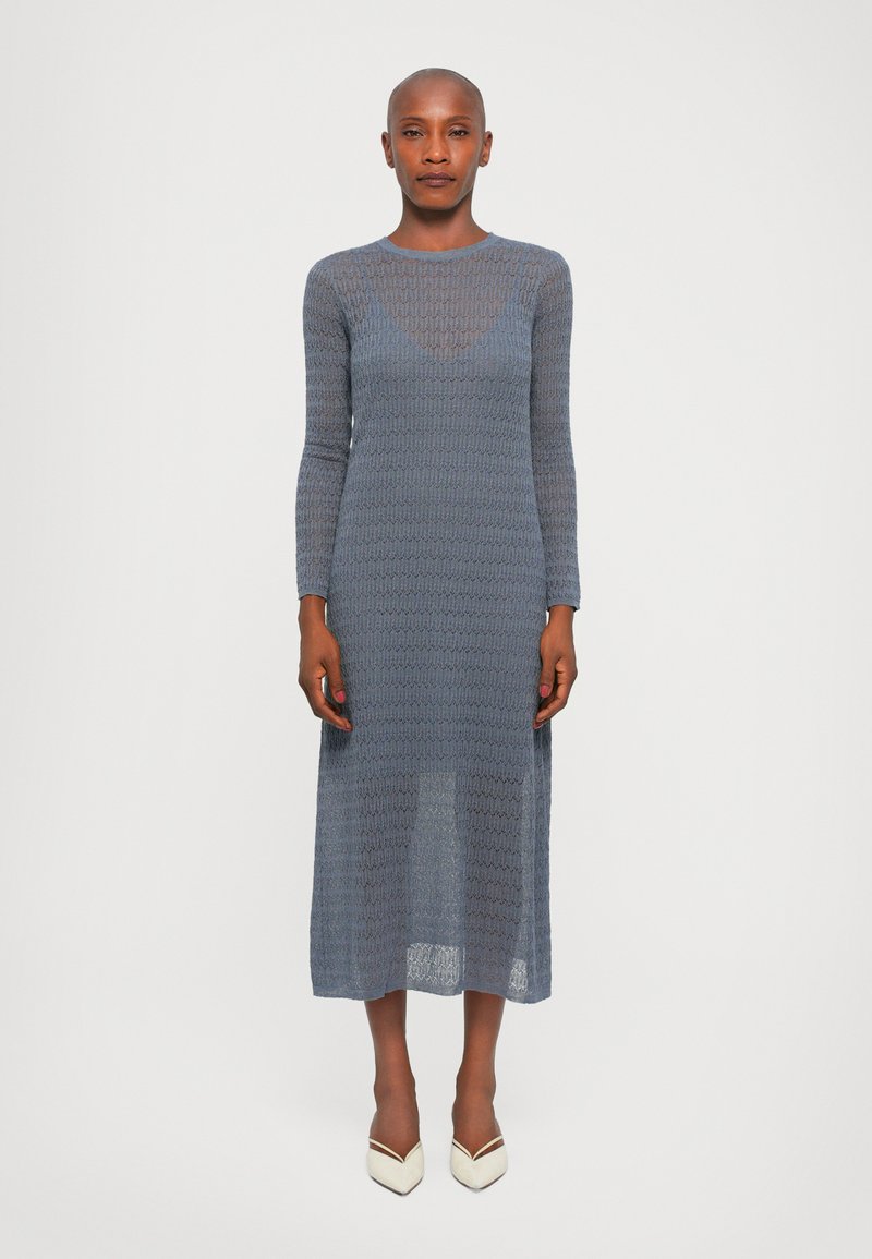 Rochie tricotată, cu mâneci lungi, de culoare albastră, cu un model texturat, până la jumătatea gambei, dintr-un material semi-transparent, asortată cu pantofi bej cu vârful ascuțit.