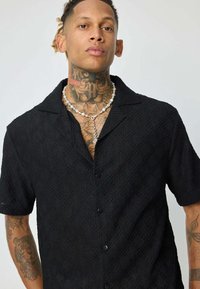 Chemise noire à manches courtes avec un motif en losanges texturé, fermeture à boutons et col. Présente une coupe décontractée et un léger éclat.