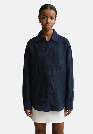 Femme à la peau foncée et aux cheveux attachés, portant une chemise ample bleu foncé à boutons et une mini-jupe blanche, debout devant un fond uni.