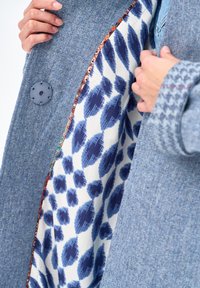 Osoba trzymająca otwarty niebieski, teksturowany płaszcz, ukazujący białą podszewkę z nieregularnymi niebieskimi plamkami oraz kontrastową lamówkę z wzorem.