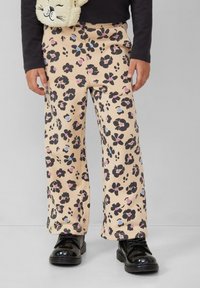 Kind dat beige broeken draagt met een zwart, roze en blauwe bloemenprint, een zwart langemouwen shirt en zwarte veterschoenen op een grijze achtergrond.