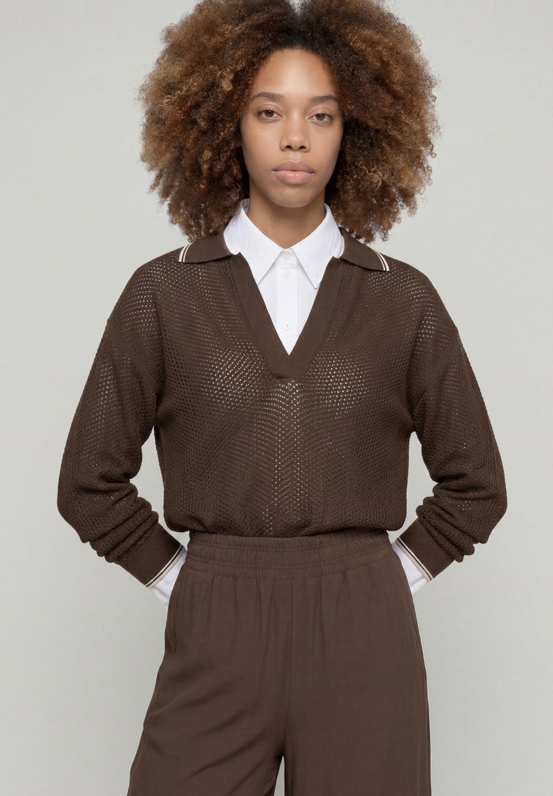 Jeune femme aux cheveux bouclés portant un pull en maille marron par-dessus une chemise blanche à col et un pantalon marron assorti, debout les mains sur les hanches.
