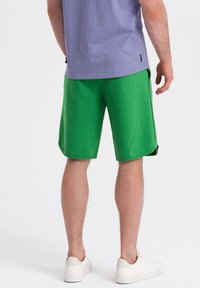 Shorts de sport verts en tissu doux, avec une texture lisse et des poches latérales, accompagnés d'un t-shirt violet clair et de chaussures blanches.