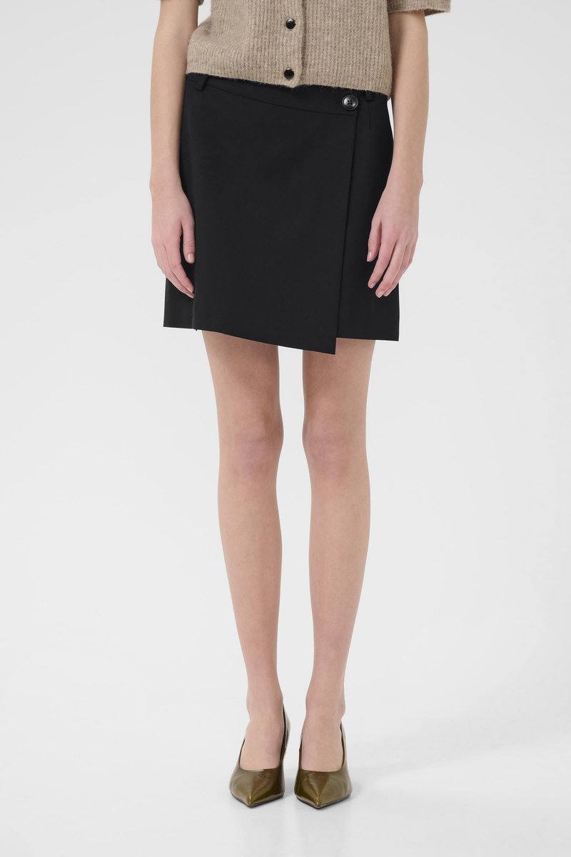 Black wrap skirt with an asymmetrical hem, featuring a side button closure. Material appears smooth; paired with pointed-toe green heels.