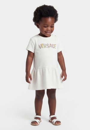 Kleines Mädchen lächelt, trägt ein weißes Kleid mit floralem Muster und weiße Sandalen, steht vor einem schlichten hellen Hintergrund.