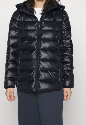 Vrouw draagt een zwarte, glanzende, gewatteerde jas met een ritssluiting aan de voorkant en capuchon, gecombineerd met donkere broek tegen een effen achtergrond.