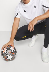 Sportowiec w białej koszulce z czarnymi akcentami klęczy obok piłki nożnej w czarno-białe wzory, ubrany w czarne spodnie i białe korki.