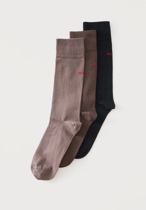 Trois paires de chaussettes type crew en beige, marron et noir, chacune avec un petit logo rouge sur le côté.