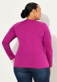 Langärmliges Shirt in lebhaftem Lila, aus weichem Stoff gefertigt. Es hat einen runden Ausschnitt und einen figurbetonten Schnitt, kombiniert mit Jeans.