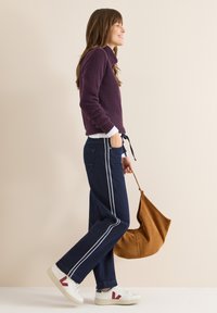 Suéter morado texturizado, pantalones anchos azul marino con rayas blancas a los lados, zapatillas blancas con detalles en rojo y bolso de hombro de ante marrón.