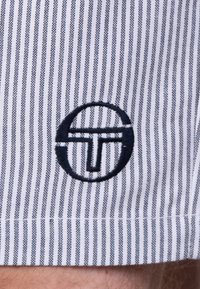 Stoff mit weißen und marineblauen Streifen, der ein kreisförmiges Logo mit einem abstrakten "T"-Design in marineblau bestickt zeigt.