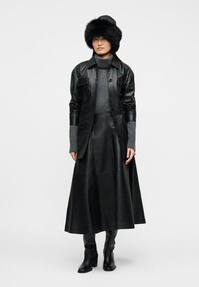 Γυναίκα που φοράει μαύρο γούνινο καπέλο, μαύρο δερμάτινο μπουφάν, γκρι πουλόβερ με ζιβάγκο, μαύρη δερμάτινη μίντι φούστα και μαύρες μπότες με τακούνι, στέκεται μπροστά σε απλό φόντο.