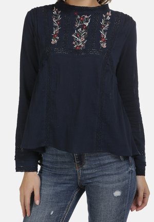 Granatowa bluzka z długim rękawem z kwiatowym haftem na klatce piersiowej, zdobiona koronkowymi detalami, zestawiona z przetartymi niebieskimi dżinsami.