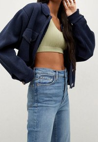 Nainen, jolla on päällään vaaleanvihreä ribbineuleinen crop-top, korkeavyötäröiset vaaleansiniset farkut ja ylisuuria tummansinisiä takkeja pitkillä hihoilla.