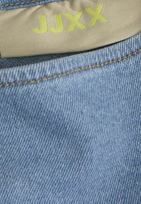 Vaate denim-kangasta vaaleansinisenä, jossa on sileä pinta. Sen vyötärönauhaan on ommeltu keltainen brodeerattu brändilippu.