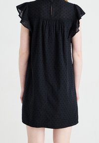 Musta mekko, joka on valmistettu kevyestä kankaasta, jossa on teksturoitu pistekuvio. Varustettu röyhelöhihoilla ja takana nappikiinnitys.