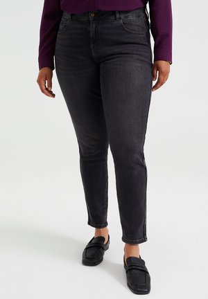 Jeans negros ajustados con una textura suave, cintura media y cinco bolsillos, combinados con mocasines negros. Fondo liso.