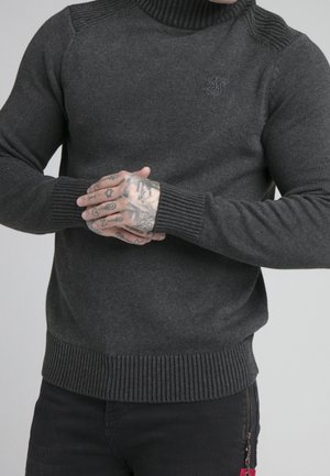 Homme portant un pull à col roulé gris foncé et un pantalon noir, les mains jointes montrant plusieurs tatouages sur les doigts et le dos de la main.