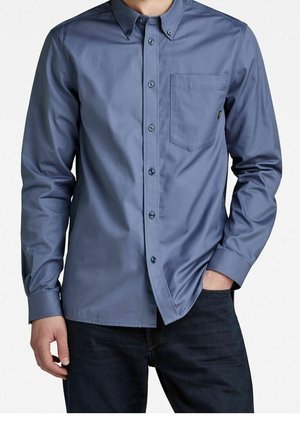 Homme portant une chemise bleue à manches longues avec poche poitrine et pantalon foncé, une main dans la poche et l'autre reposant sur le côté.