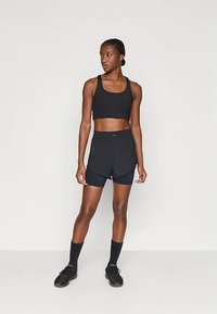 Aktiivne naine seisab, seljas must spordipäevik, kihilised mustad jooksuhõlmad, mustad sokid ja mustad tossud, kahvatuhalles taustal.