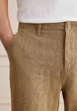 Lähivõte inimesest, kes kannab pruune kalasaba mustriga pükse, millel on nupp ja vööaas, käsi vasakus taskus, valget tippu kandes.