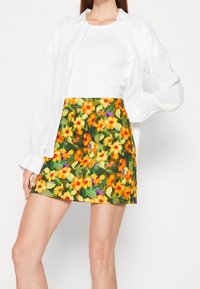 Falda mini floral con vibrantes flores amarillas y naranjas sobre un fondo verde; combinada con una camiseta blanca ajustada y una camisa blanca.