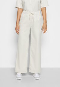 Personne portant un pantalon ample blanc cassé avec une taille élastique et un cordon, associé à des baskets blanches et une chemise boutonnée à manches courtes.