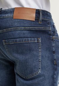 Takap pocket dan pinggang celana denim biru dengan label kulit cokelat bertuliskan "2Y Premium," dipadukan dengan kemeja putih yang dimasukkan.