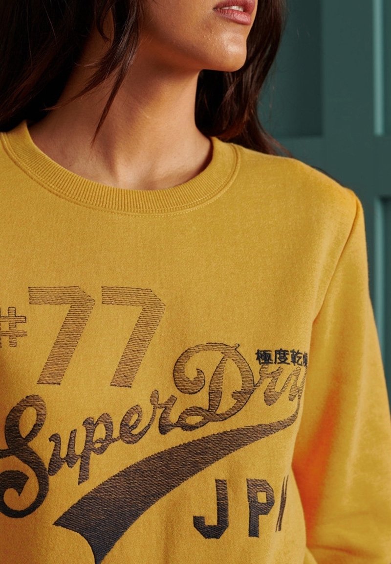 Femme portant un sweat-shirt jaune moutarde avec le texte "#77 SuperDry JP/" et des caractères japonais sur la poitrine, sur fond vert.