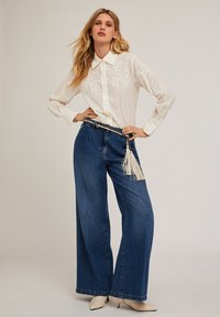 Chemise blanche à manches longues avec des détails brodés, associée à un jean bleu à jambe large et une ceinture à franges. Des chaussures beiges à bout pointu complètent le look.