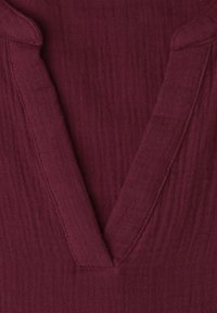 Gros plan sur un col en V profond d'un vêtement en tissu texturé et doux de couleur bordeaux foncé.