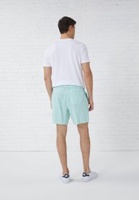 Men's white polo shirt above light green and white striped shorts, with a back pocket and solid color shoes. Set against a white brick wall.