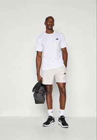 Weißes Sport-T-Shirt mit schwarzem Logo, hell gemusterte Shorts, schwarze Sneakers, weiße Socken und eine dunkle Sporttasche, stehend vor einem neutralen Hintergrund.
