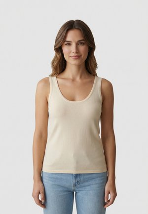 Femme aux cheveux bruns mi-longs portant un haut sans manches beige clair et un jean bleu clair, debout devant un fond blanc uni.