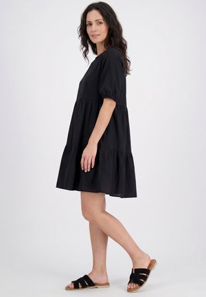 Femme en robe noire ample à étages et sandales noires ouvertes, debout de profil sur un fond clair uni.