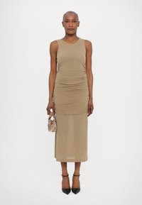 Rochie tricotată, fără mâneci, de culoare bej, cu detalii pliate pe laterale, ajungând până la jumătatea gambelor. Asortată cu o poșetă mică, cu model, și pantofi cu toc negri.