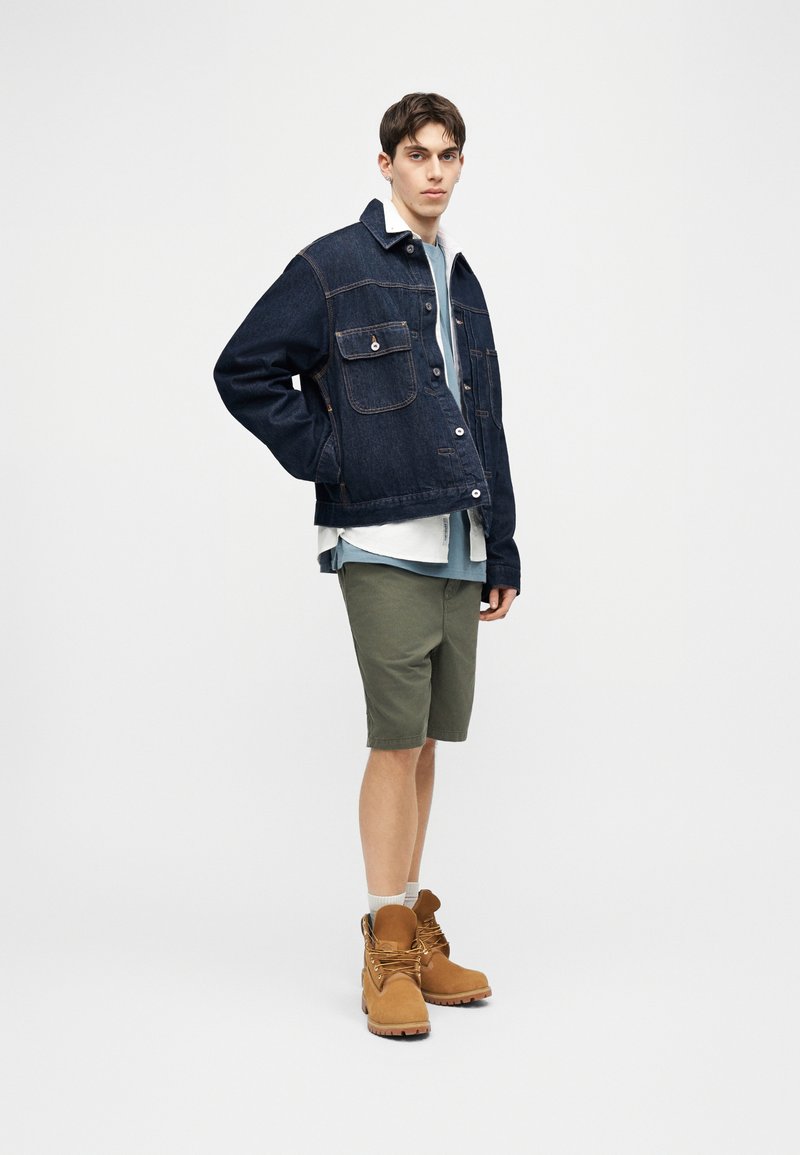 Junger Mann mit dunkler Jeansjacke, blauem Hemd, grünen Shorts und hellbraunen Arbeitsstiefeln, der vor einem schlichten hellen Hintergrund steht.