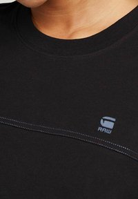 Černé tričko s krátkým rukávem z bavlny s kulatým výstřihem, s modrým kontrastním šitím a malým logem "RAW" na hrudi.