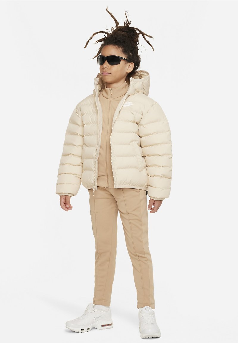 Chaqueta de plumas crema con secciones acolchadas horizontales, chándal ajustado beige y zapatillas blancas. Gafas de sol oscuras completan el conjunto.