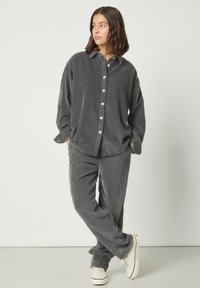 Femme debout les mains dans les poches, portant une chemise et un pantalon en velours côtelé gris foncé amples avec des baskets blanches, sur un fond uni.