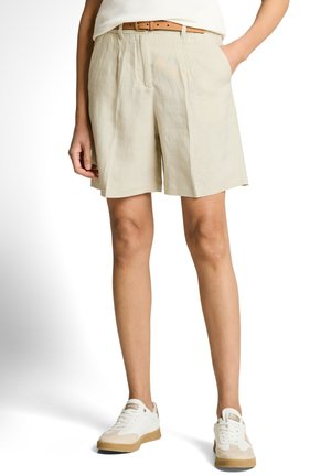 Persona que lleva puestos unos pantalones cortos plisados beige con bolsillos laterales, un cinturón marrón claro y zapatillas blancas con detalles beige sobre un fondo blanco.