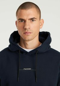 Felpa con cappuccio blu navy dotata di un cordino, con un piccolo logo stampato bianco "CHASIN'" sul davanti. Tessuto morbido e vestibilità comoda.