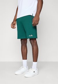 Grønne atletiske shorts lavet af et blødt stof, med sidespalter, et logo på venstre søm, og parret med hvide sneakers og sokker.