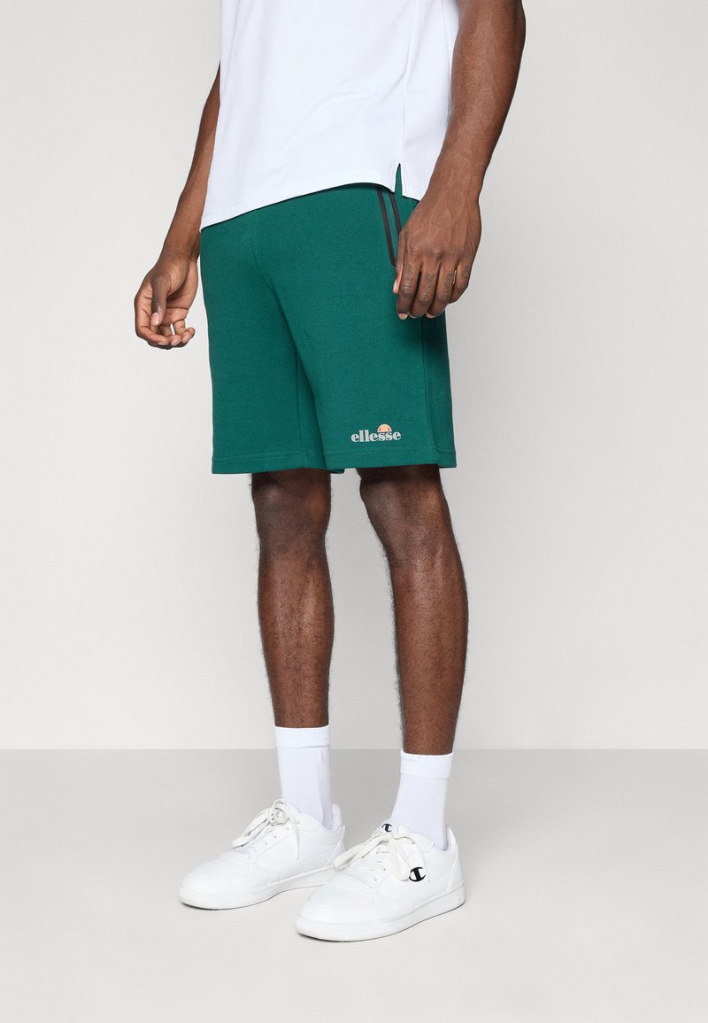 Grønne atletiske shorts lavet af et blødt stof, med sidespalter, et logo på venstre søm, og parret med hvide sneakers og sokker.
