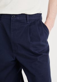 Pantalon bleu marine sur mesure avec plis, doté d'une fermeture à bouton, de poches avant et d'une texture de tissu lisse.